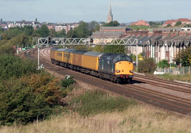 A blue diesel locomotive hauling a line of yellow test coaches along the main railway line near Hasland, with terraced houses beside the tracks and Chesterfield's Crooked Spire on the skyline.