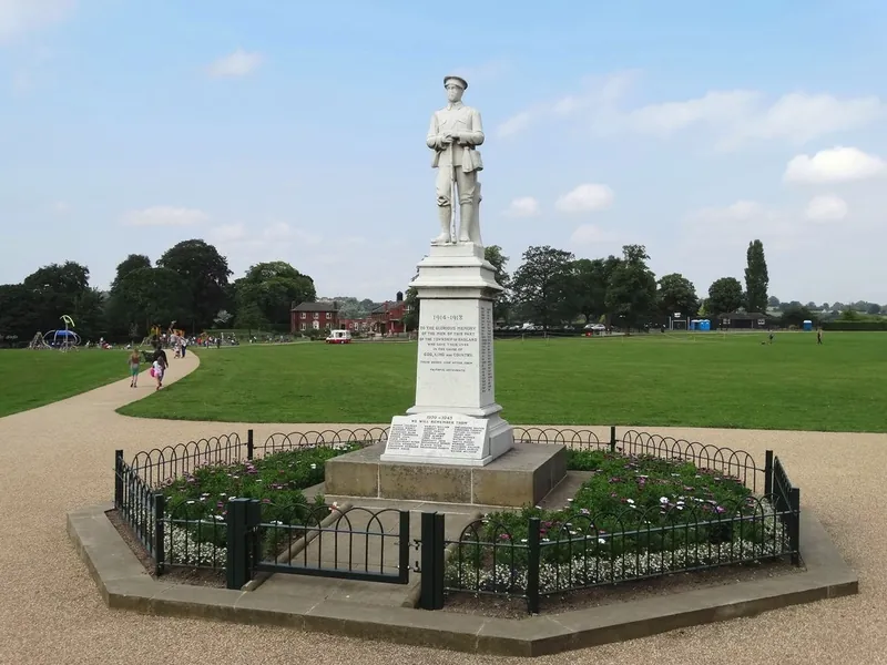 Hasland War Memorial: a white stone statue of a First World War soldier on a tall plinth inscribed 1914-1918 and 1939-1945, inside a low railed flower bed, with the open lawns and trees of Eastwood Park behind under a blue sky.